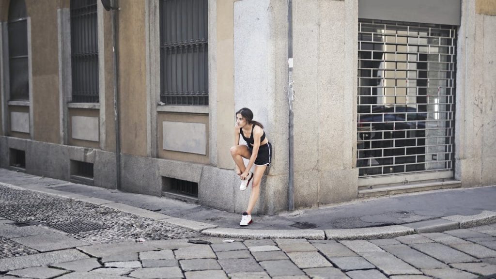 Slim female runner in activewear standing on corner of street and tying shoelaces on sneakers while leaning on stone wall of building and looking away